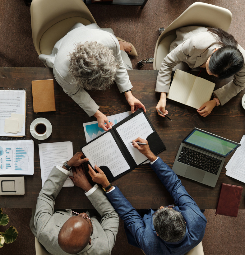 Group of middle aged multiethnic business professionals collaborating around table, reviewing documents and using laptop, top view showing teamwork and corporate meeting environment